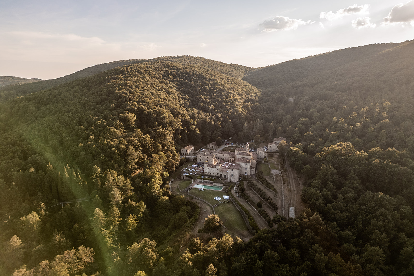 Aerial golden-hour view of SPAO Borgo San Pietro nestled in a forested Umbrian valley, with stone houses, terracotta roofs, terraced gardens and a pool